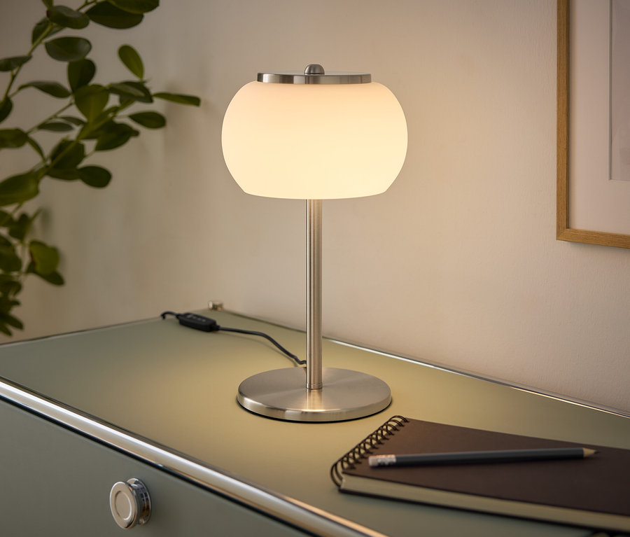 Svietiaca stolová lampa s bielym guľovým tienidlom a brúseným kovovým podstavcom stojí na svetlozelenej komode. Vedľa nej leží zápisník a ceruzka. Vľavo je rastlina, vpravo zarámovaný obraz na stene.
