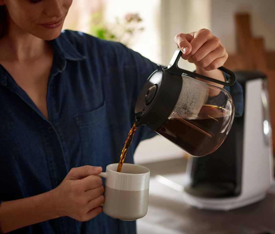 Žena nalieva kávu z kanvice do šálky. V pozadí je vidieť kávovar na prekvapkávanú kávu »Let's Brew«.