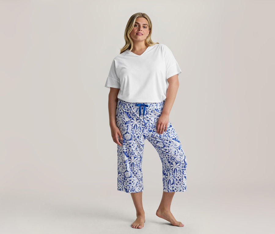 Žena stojí, oblečená v bielom tričku s V-výstrihom a modro-bielych vzorovaných 3/4-Pyjama-Set nohaviciach s modrou šnúrkou, bosá.