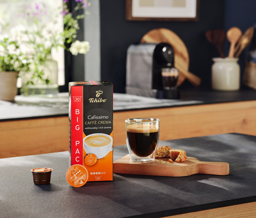 Balenie kapsúl Cafissimo Caffe Crema, pohár s kávou a kúsky sušienok na stole. V pozadí kávovar Cafissimo pure+ black.
