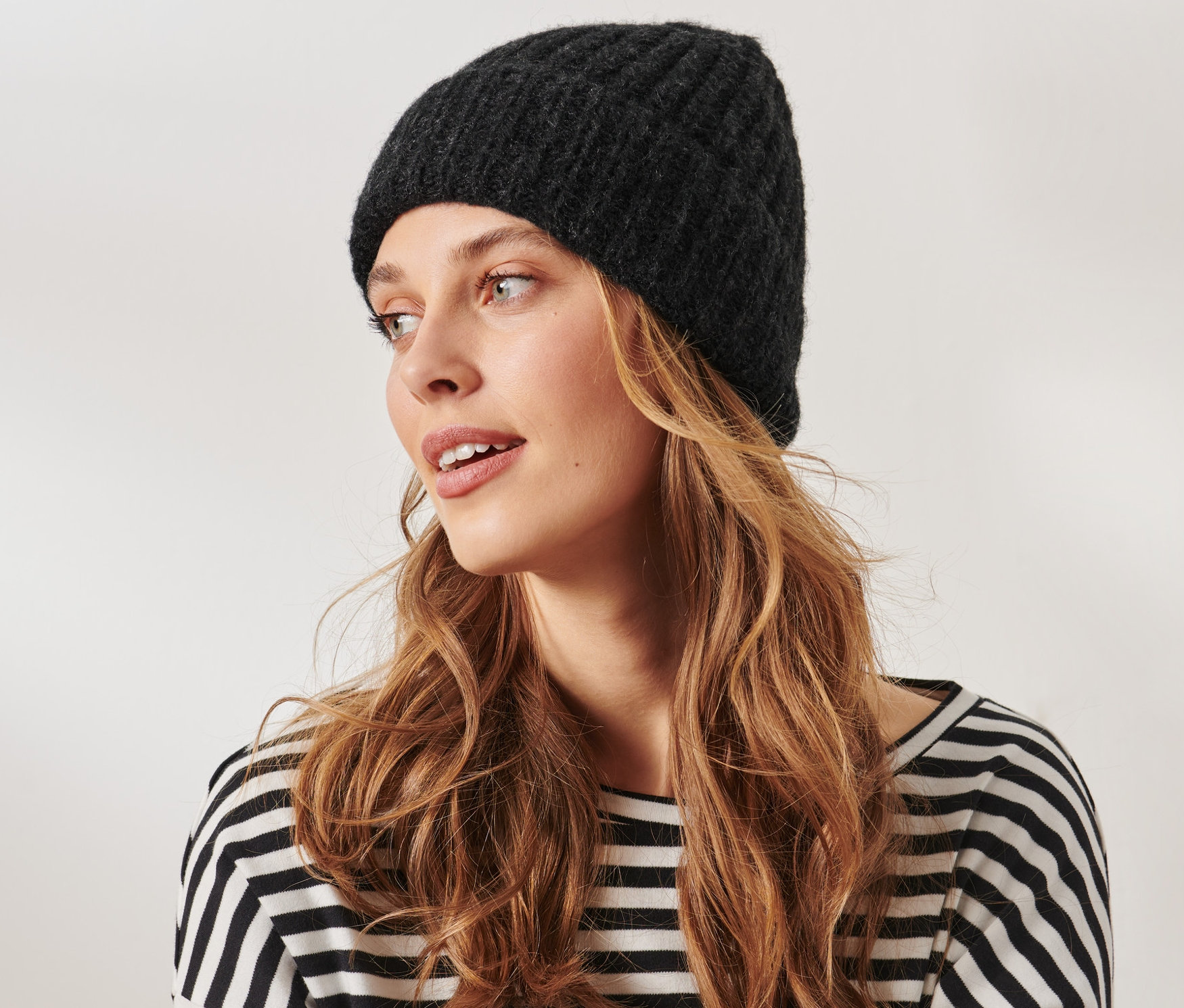 Žena s dlhými vlnitými vlasmi a v pletenej čiapke s vlnou, antracitová, sa pozerá doprava.