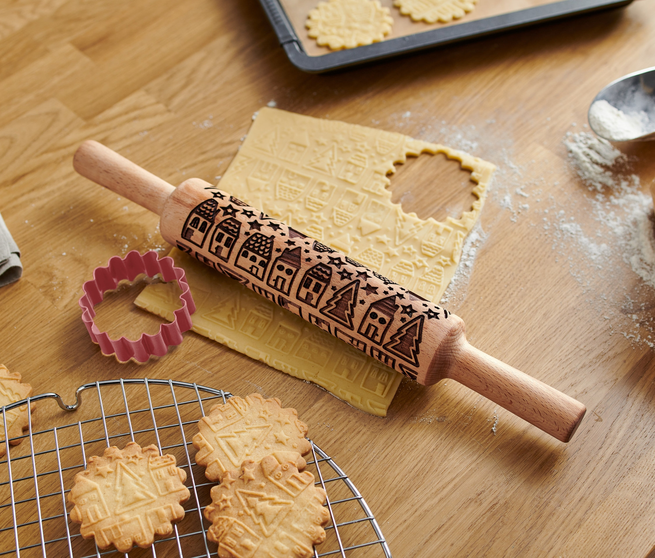 Na drevenom stole leží valček na cesto s motívom, vykrajovátko a upečené sušienky.