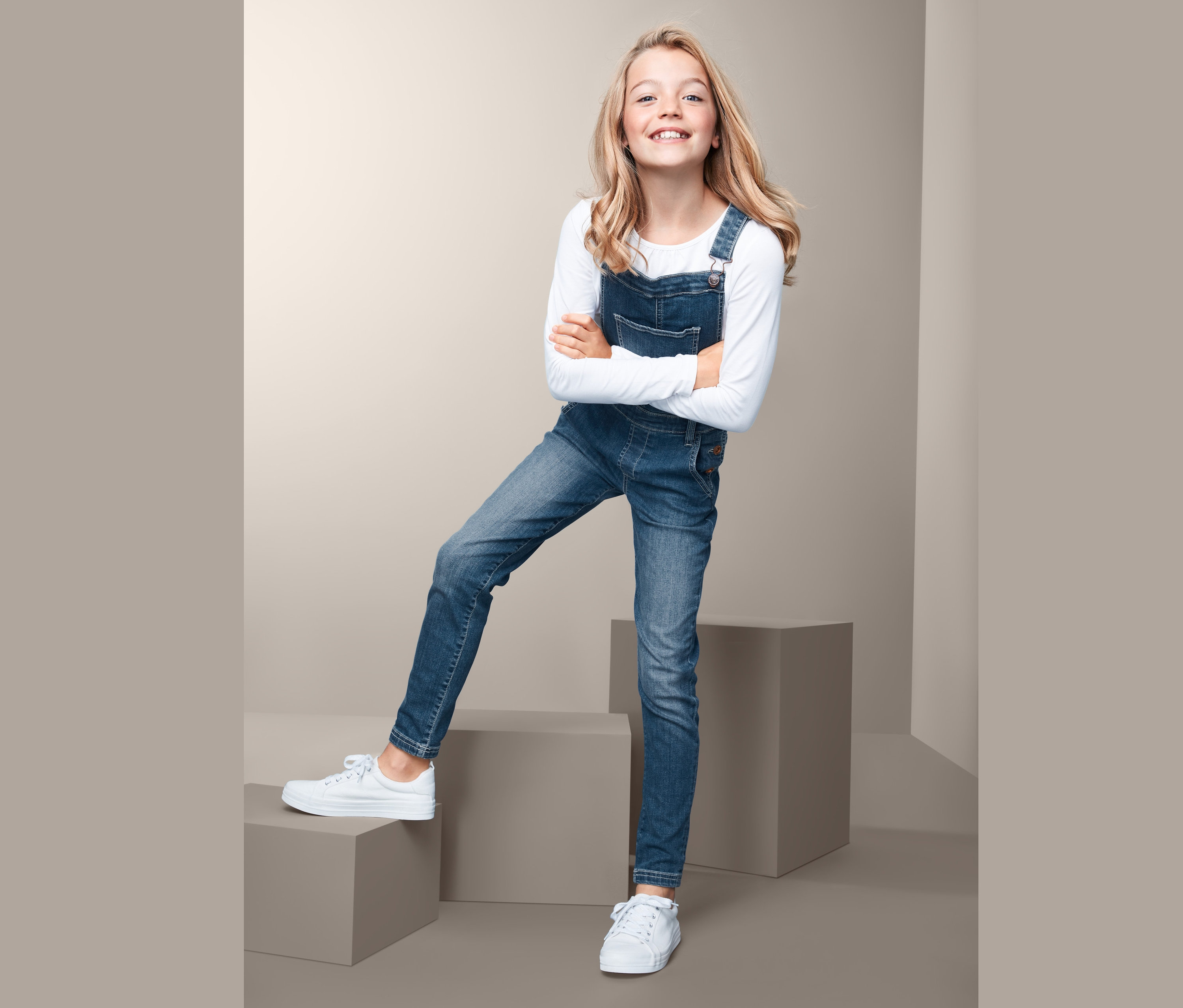 Usmievajúce sa dievča pózuje so skríženými rukami a má na sebe džínsy s náprsenkou a biele tenisky.