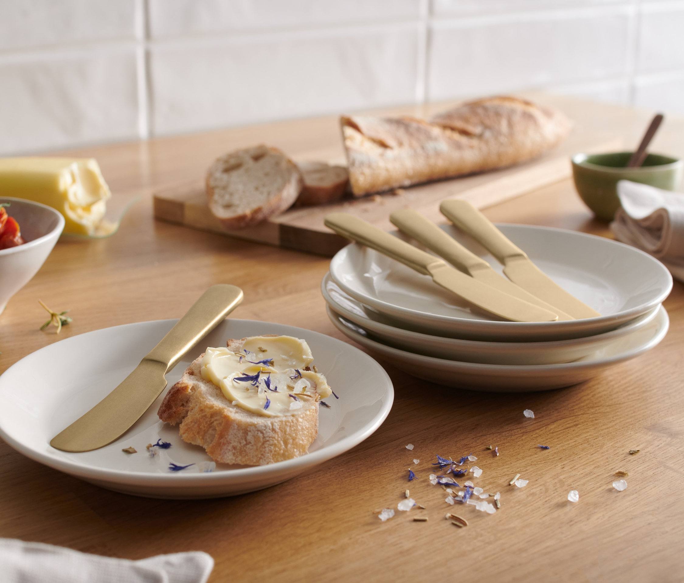 Na stole leží chlieb s maslom a nôž na maslo, vedľa sú 4 nože na maslo na tanieroch.