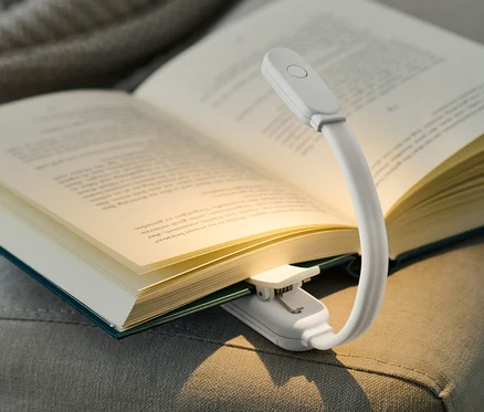 Otvorená kniha s LED lampou na čítanie, umiestnená na šedej pohovke.