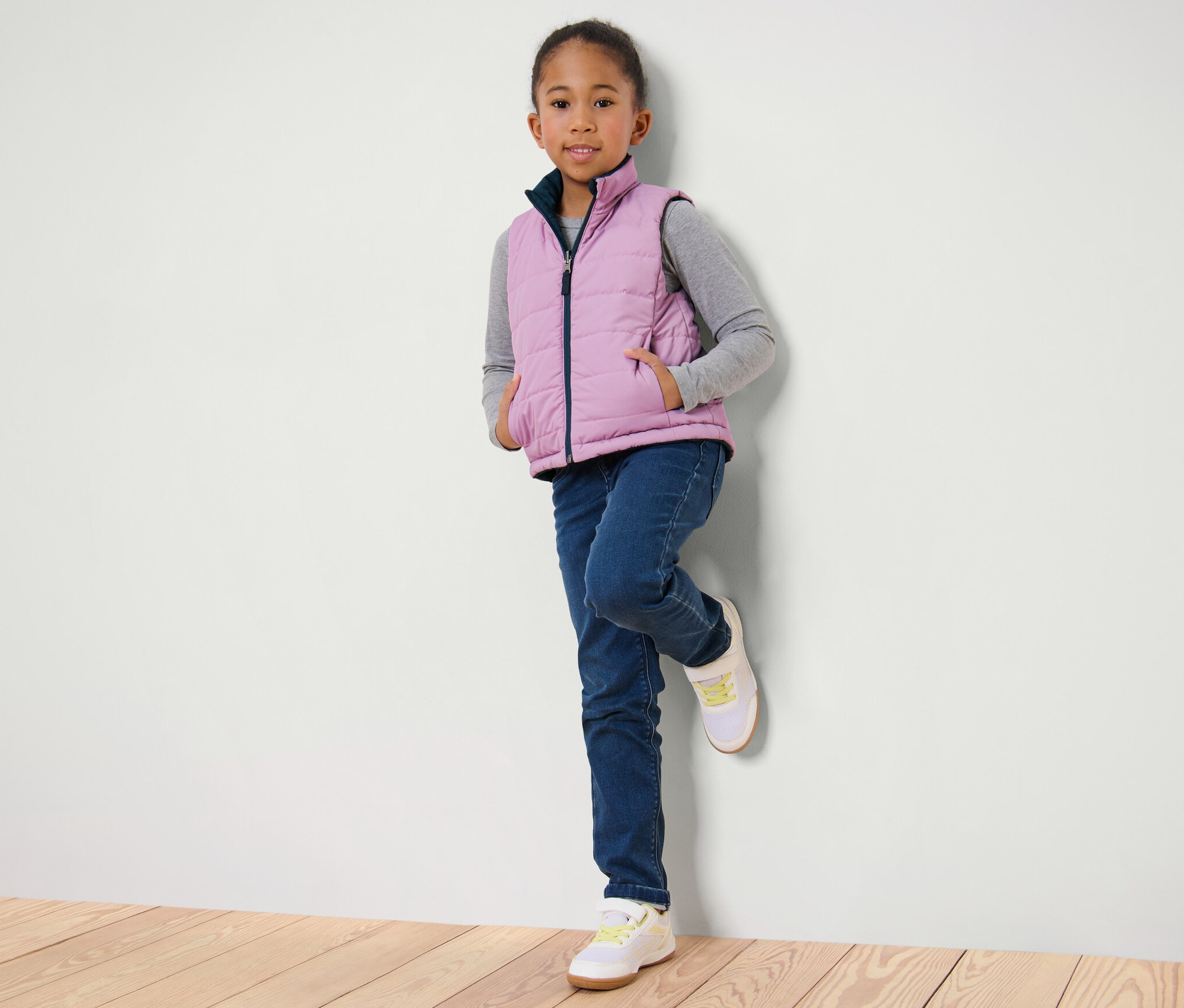 Dievča opreté o stenu má na sebe detskú obojstrannú prešívanú vestu, džínsy a tenisky.