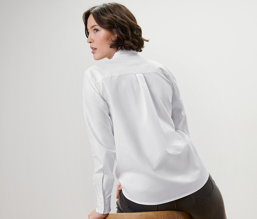 Žena sedí bokom na drevenej stoličke a má na sebe bielu blúzku s nariaseným detailom.