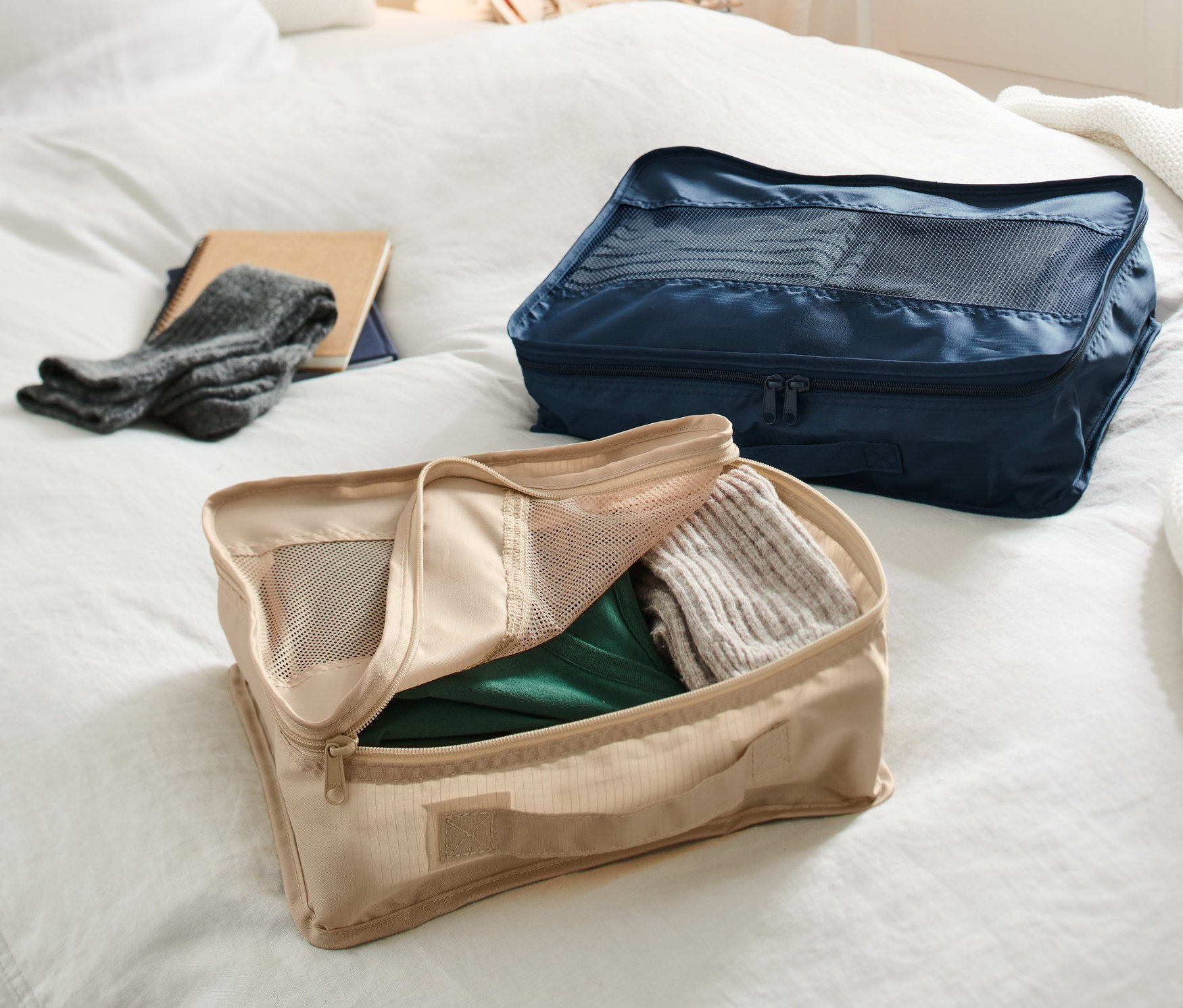 Dva cestovné organizéry ležia na posteli. Béžový organizér je otvorený a plný oblečenia, zatiaľ čo tmavomodrý organizér je zatvorený.