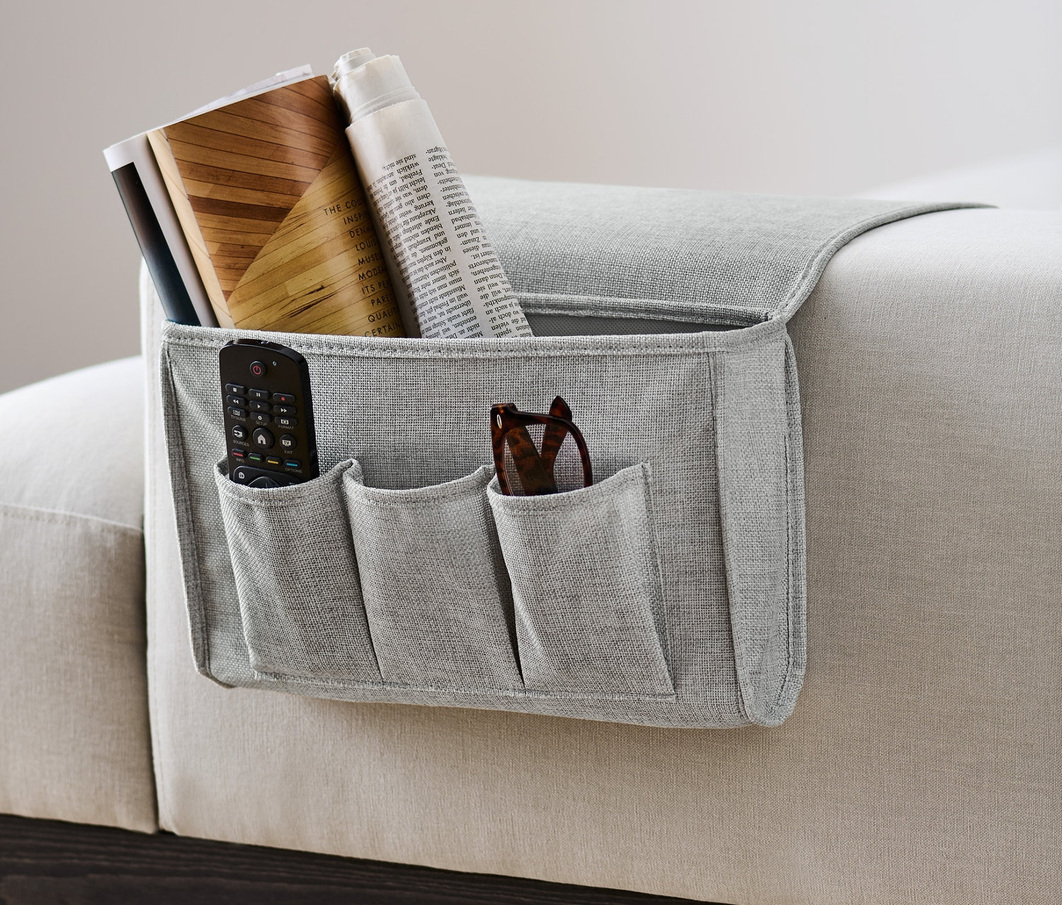 Sivý organizér na posteľ a pohovku visí na boku pohovky a obsahuje diaľkový ovládač, okuliare, knihy a noviny.