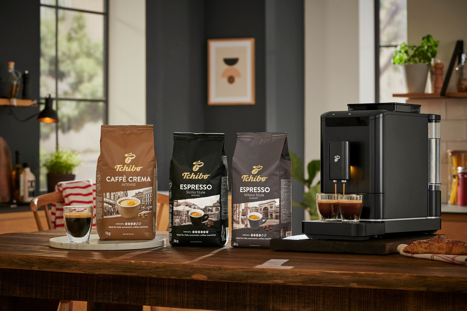 Kávovar pripravuje kávu do dvoch pohárov vedľa balení zrnkovej kávy Tchibo Caffè Crema, Espresso Sicilia Style a Espresso Milano Style.