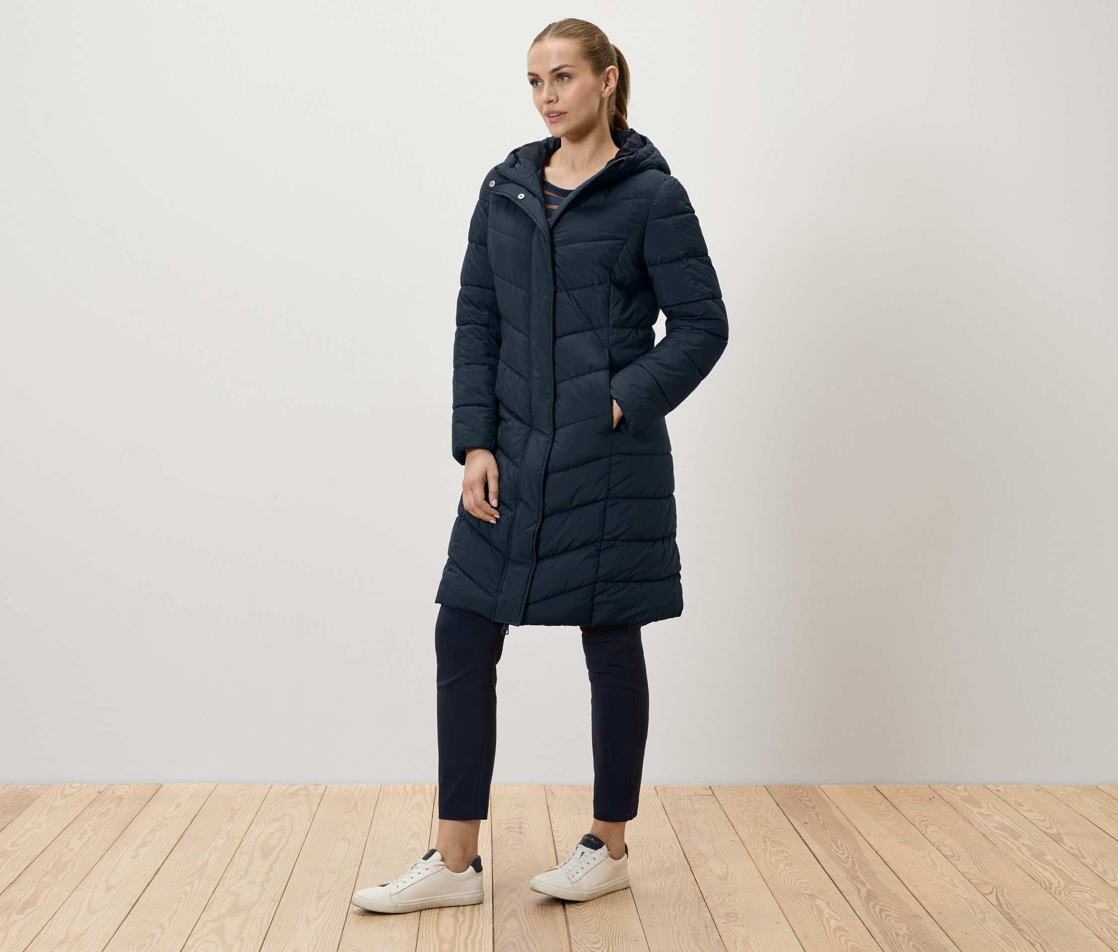 Žena pózuje v dlhom, tmavomodrom prešívanom kabáte, tmavomodrých nohaviciach a bielych teniskách.