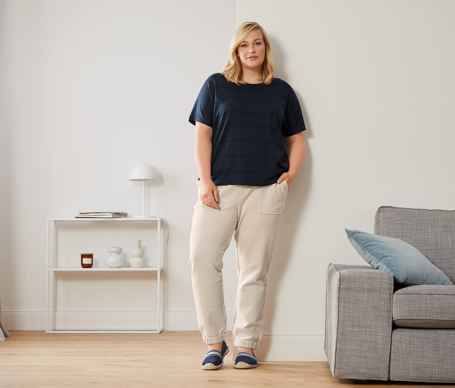 Žena opretá o stenu má na sebe tmavomodré tričko s okrúhlym výstrihom, teplákové nohavice s dĺžkou po členky a espadrilky.