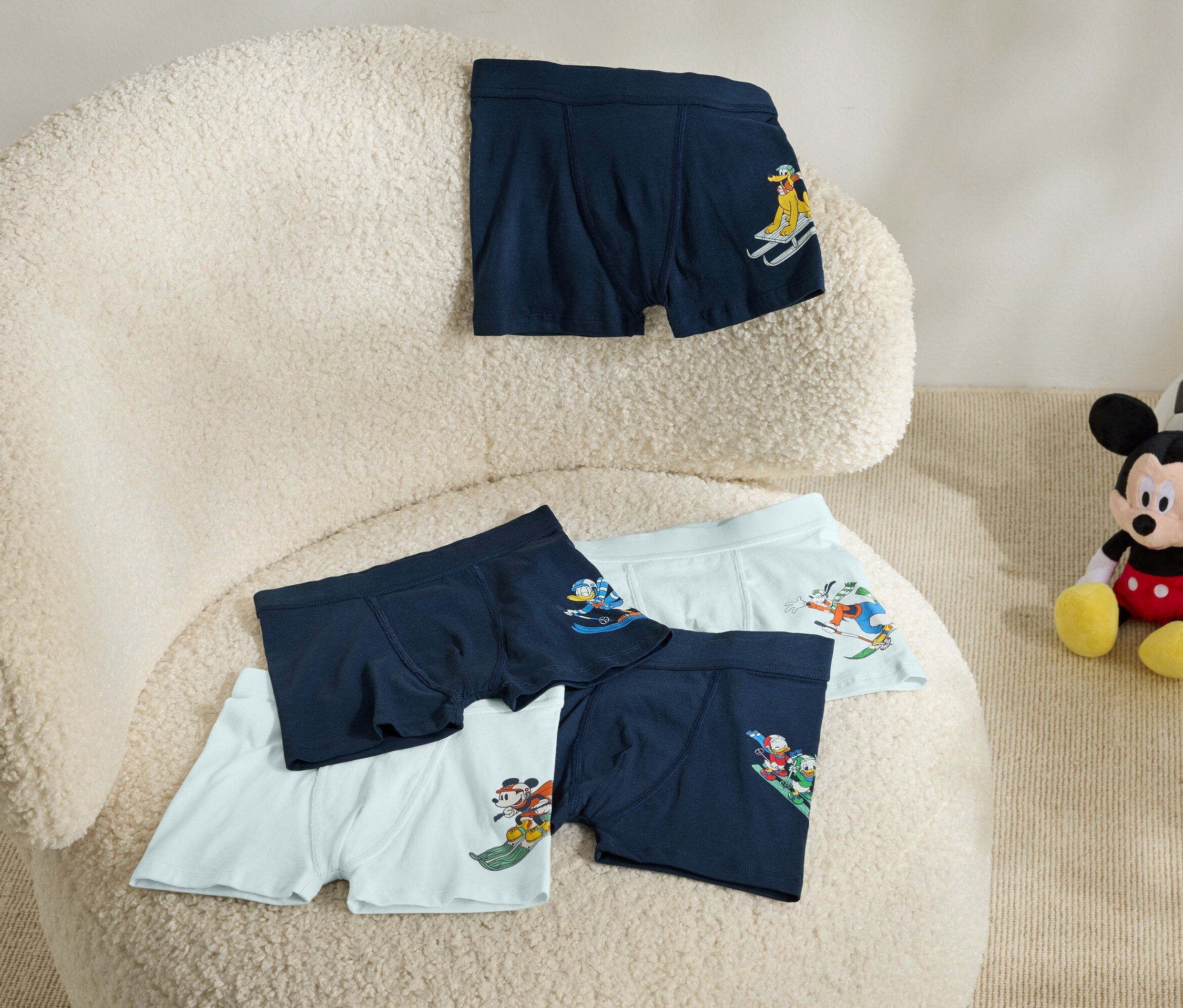 Na béžovom kresle leží päť boxeriek s potlačou. Vedľa kresla sedí plyšová hračka Mickey Mouse s futbalovou loptou.