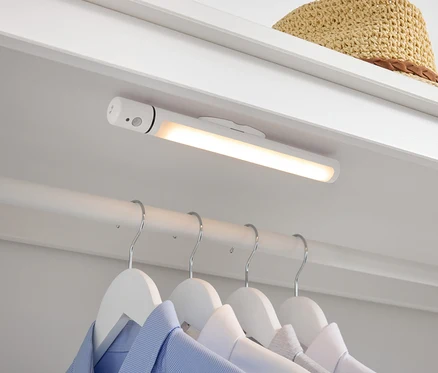 V šatníku je biela LED lampa, pod ňou visia na bielych vešiakoch košele a hore leží slamený klobúk.