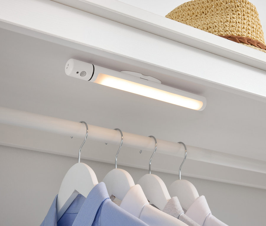 V šatníku je biela LED lampa, pod ňou visia na bielych vešiakoch košele a hore leží slamený klobúk.