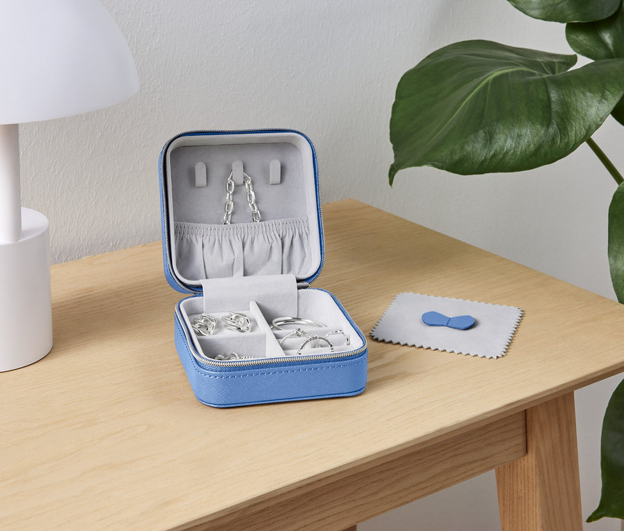 Otvorená, modrá šperkovnica so striebornými šperkami stojí na drevenom stole vedľa bielej lampy a zelenej rastliny.