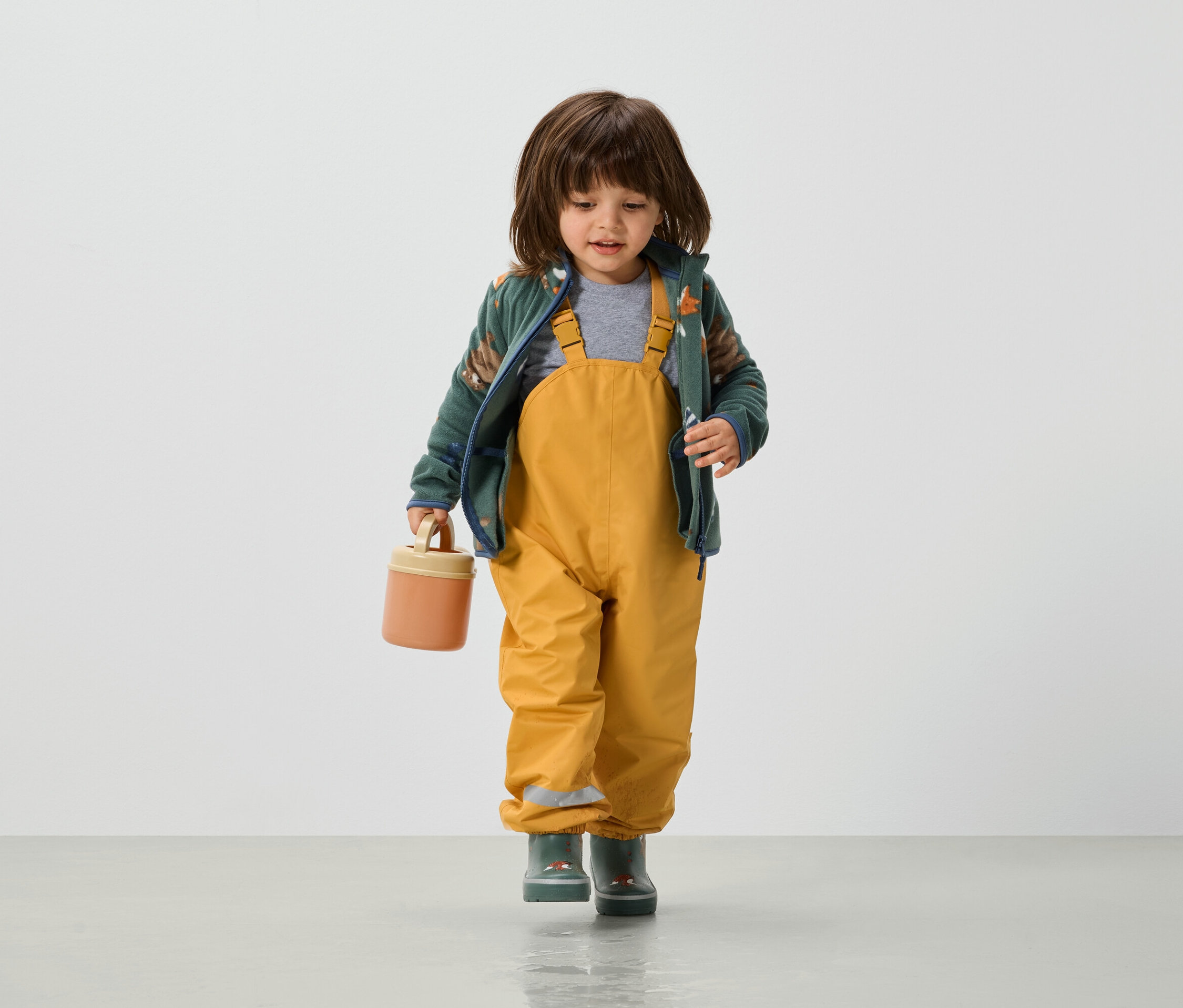 Batoľa v žltých nohaviciach s náprsenkou, zelenej bunde a gumákoch drží v ruke oranžový box na desiatu.