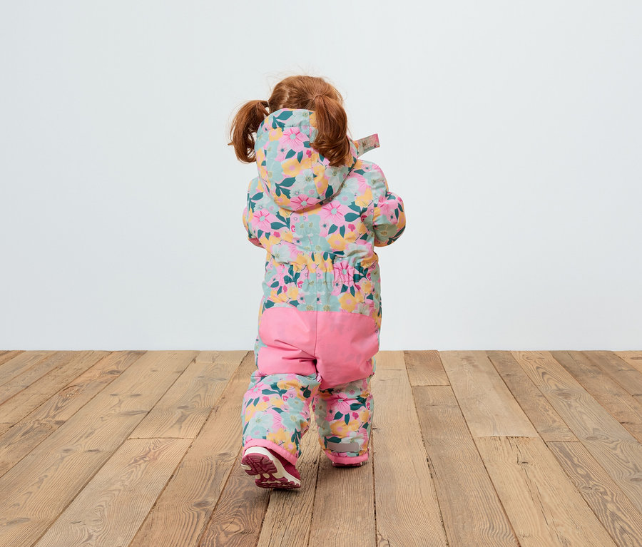 Pohľad zozadu na dieťa s ryšavými vlasmi v kvetinovom overale s kapucňou a ružovými nohavicami, ktoré stojí na drevenej podlahe.