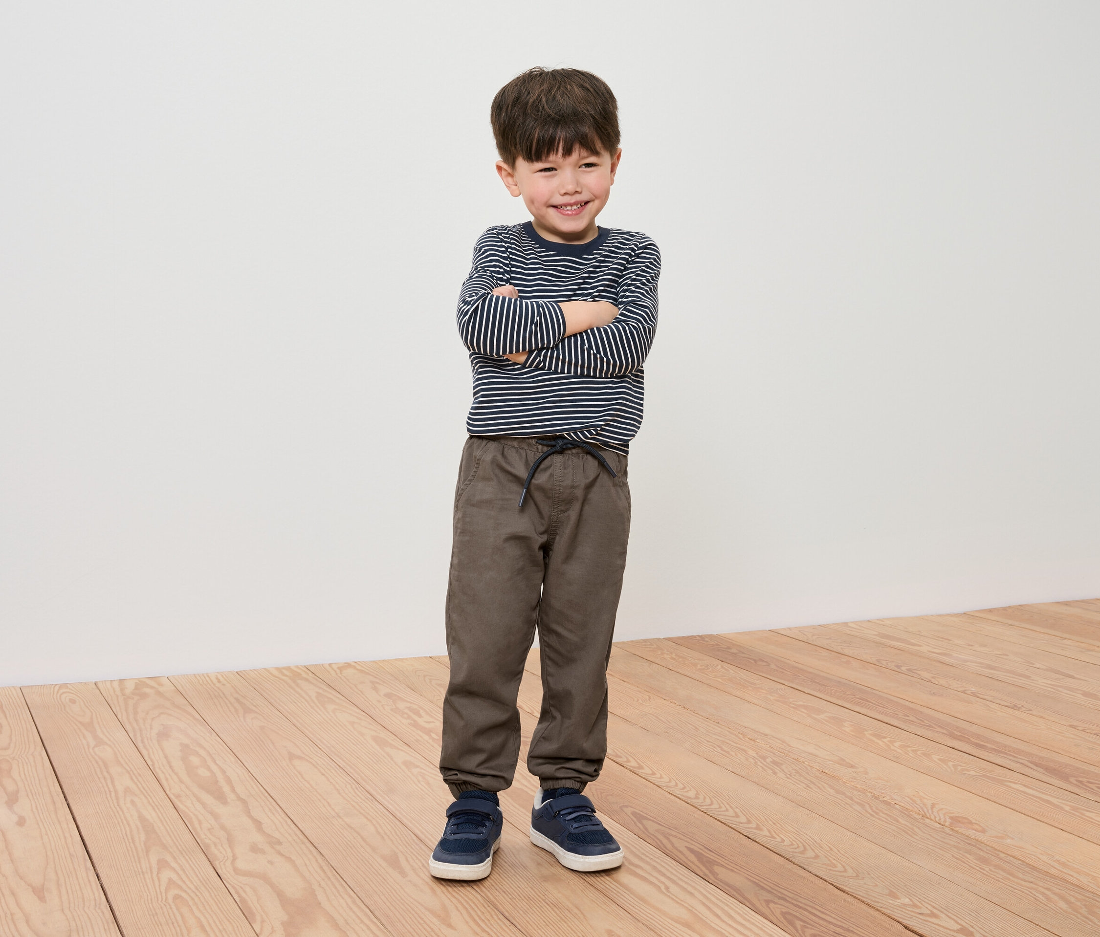 Chlapec so skríženými rukami pózuje v pruhovanom tričku, hnedých nohaviciach a modrých teniskách.