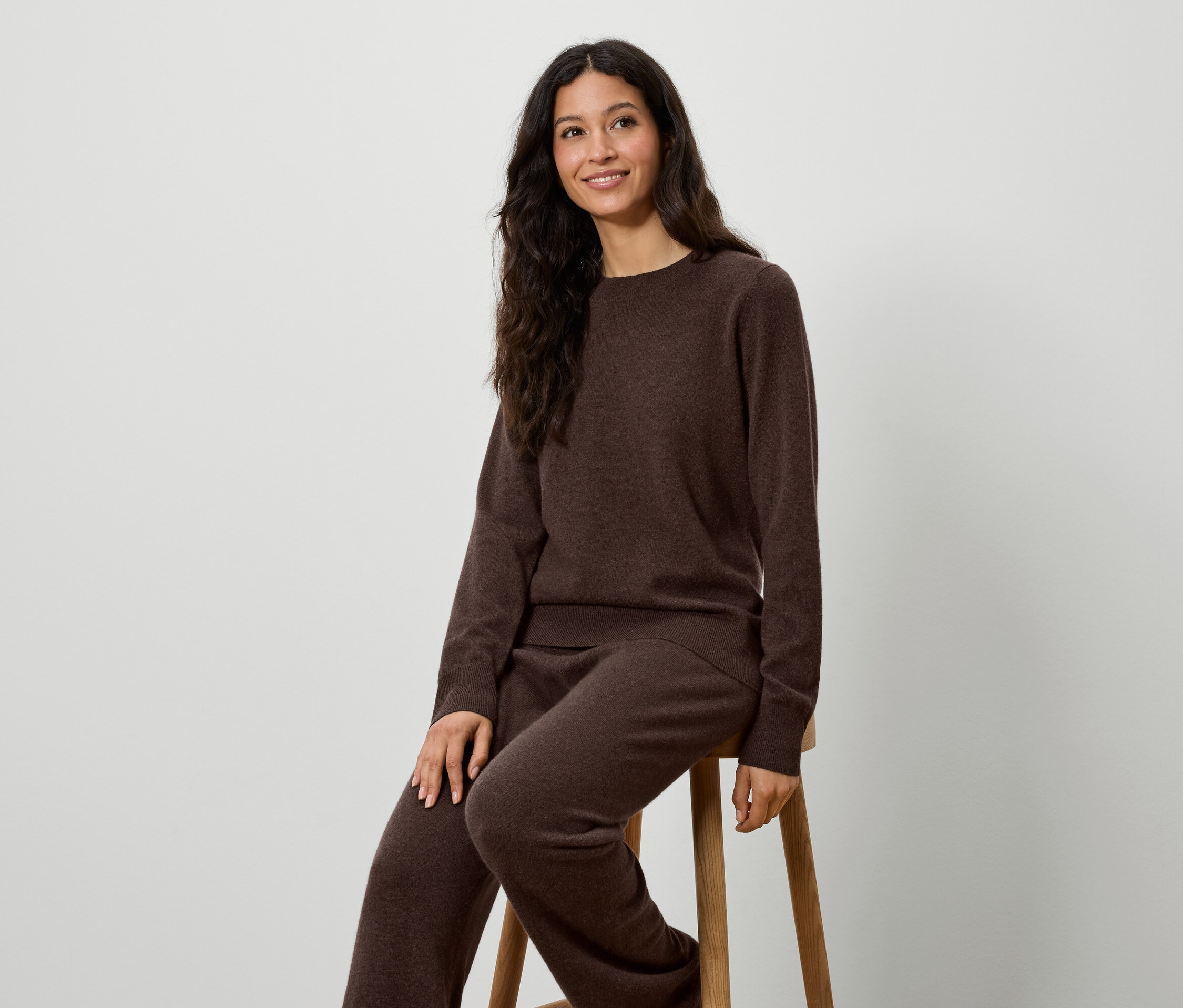 Žena sedí na stoličke a má na sebe hnedý pletený sveter a nohavice.