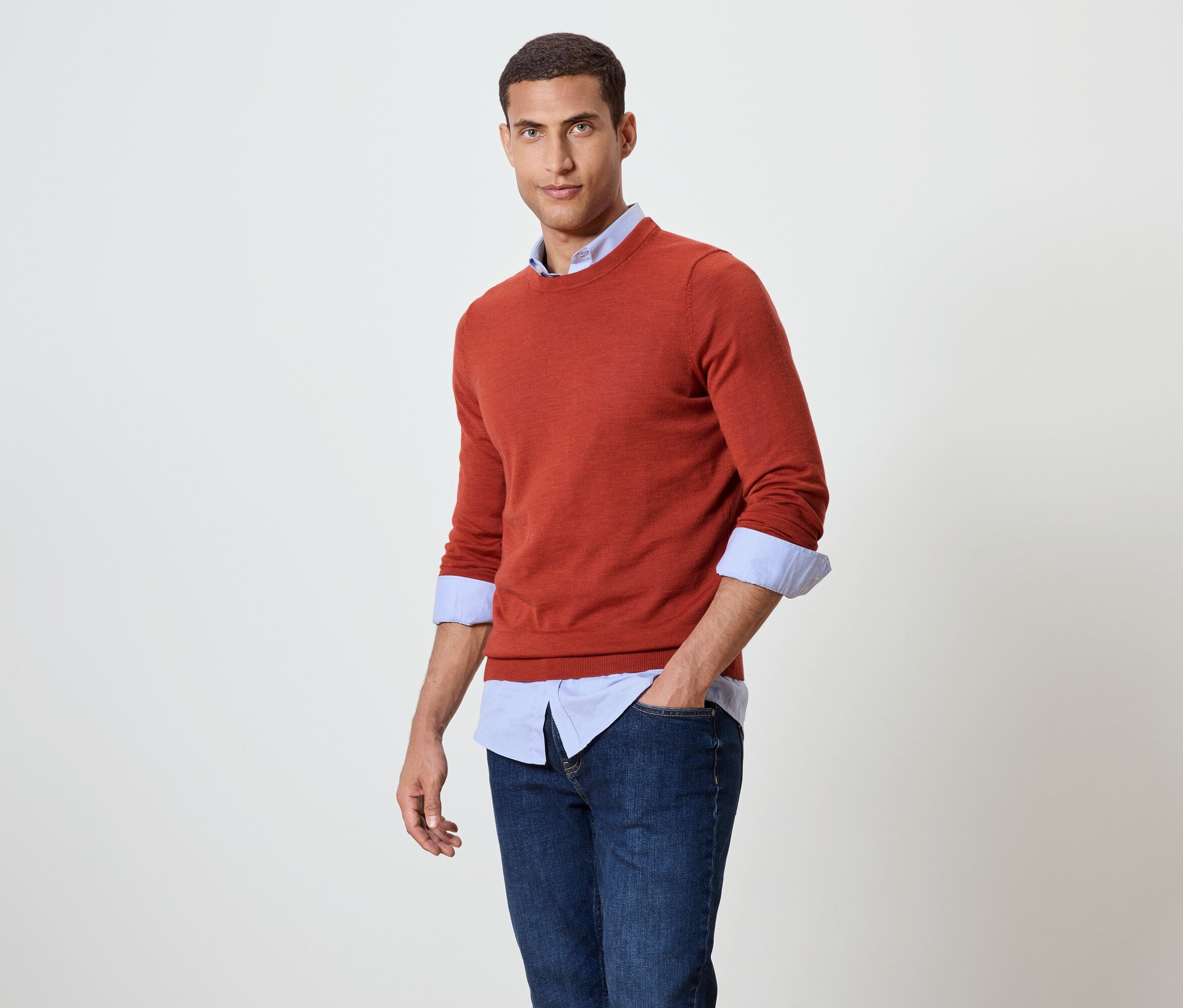 Muž pózuje v oranžovom svetri cez modrú košeľu a džínsy.