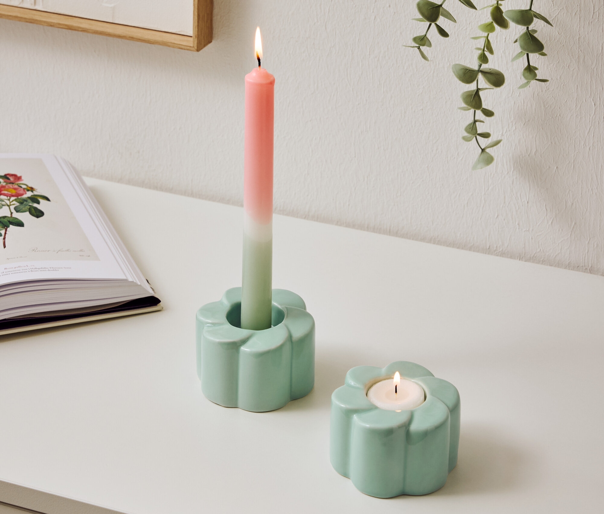 Dva svetlomodré svietniky v tvare kvetov stoja na stole, jeden s horiacou, dvojfarebnou sviečkou.