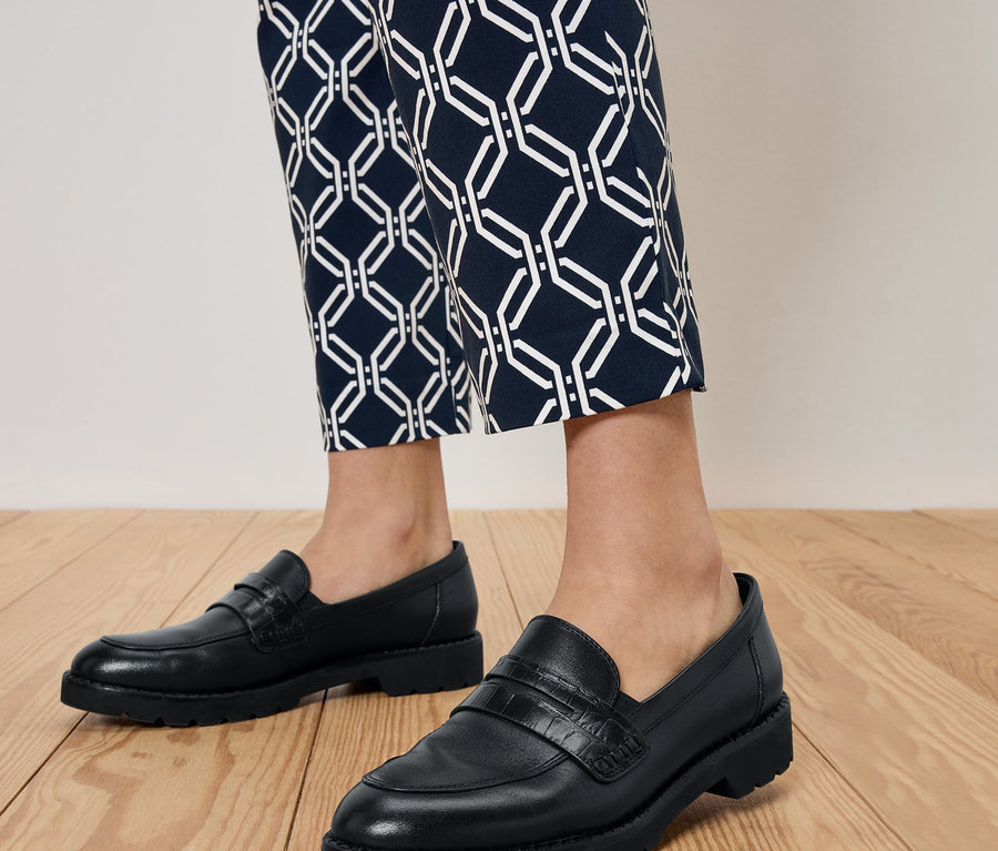 Nohy osoby v čiernych mokasínach a modrých vzorovaných nohaviciach stojacej na drevenej podlahe.