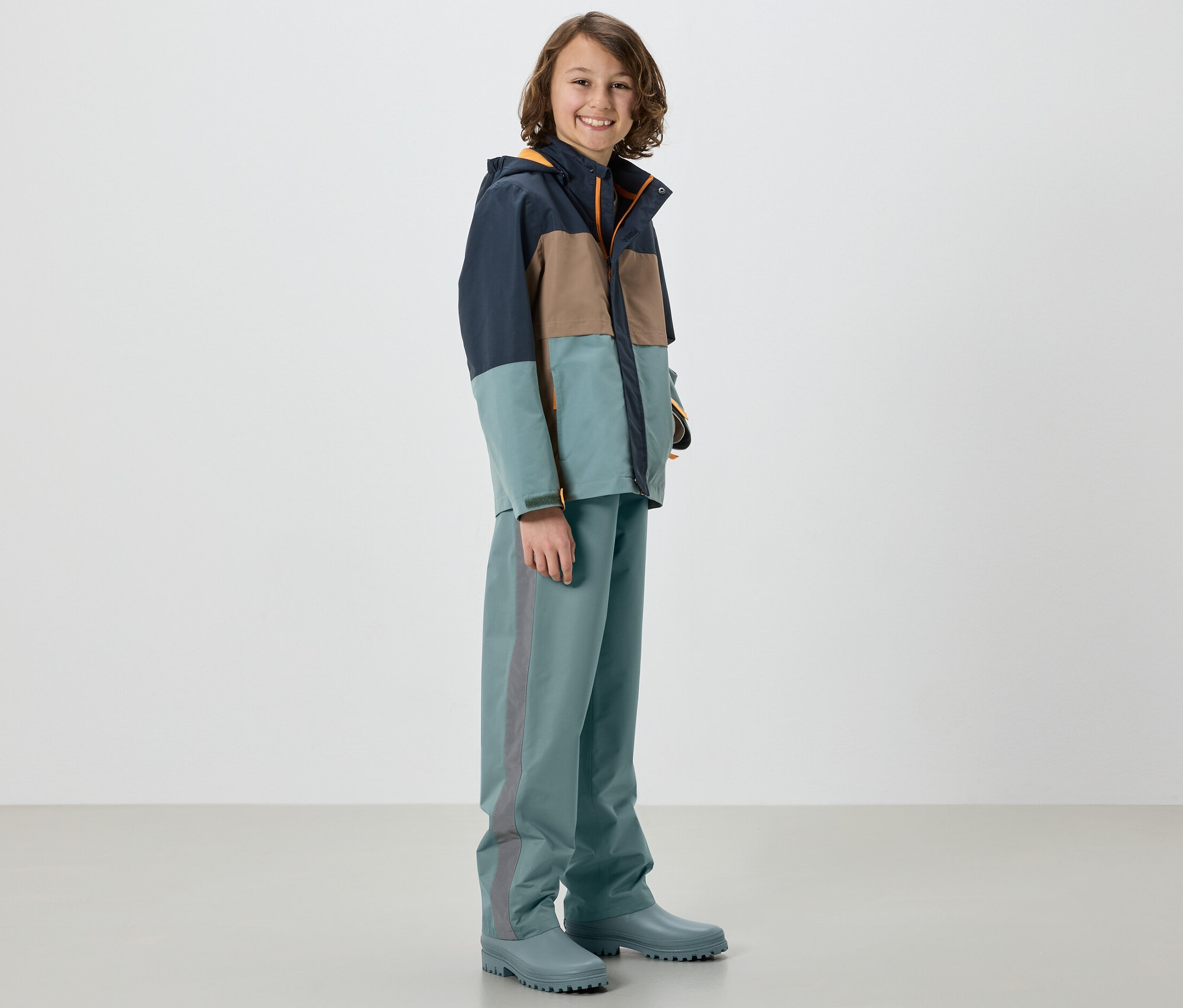 Chlapec v bunde a detských nohaviciach do dažďa, šalvia, stojí v gumákoch.