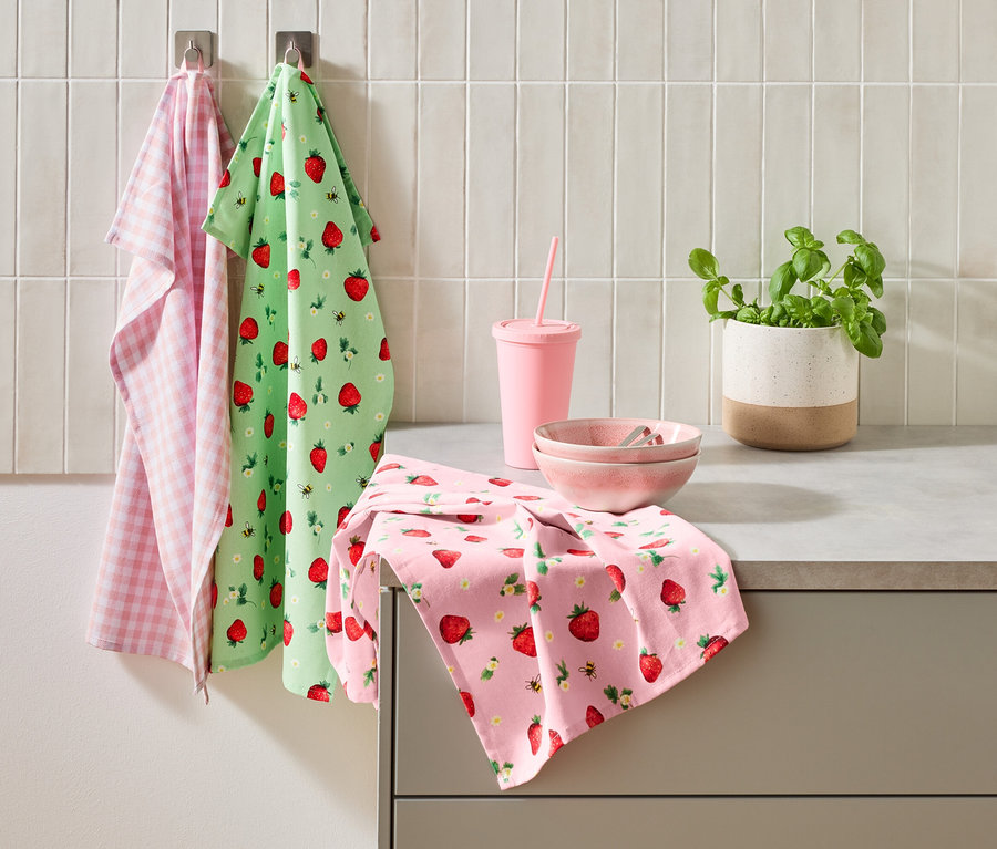 Tri kuchynské utierky, ružový pohár so slamkou, dve ružové misky a kvetináč bazalky stoja na kuchynskej linke.