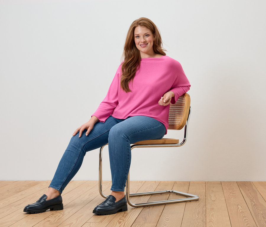 Žena sedí na stoličke a má na sebe ružový sveter, modré džínsy a čierne mokasíny.