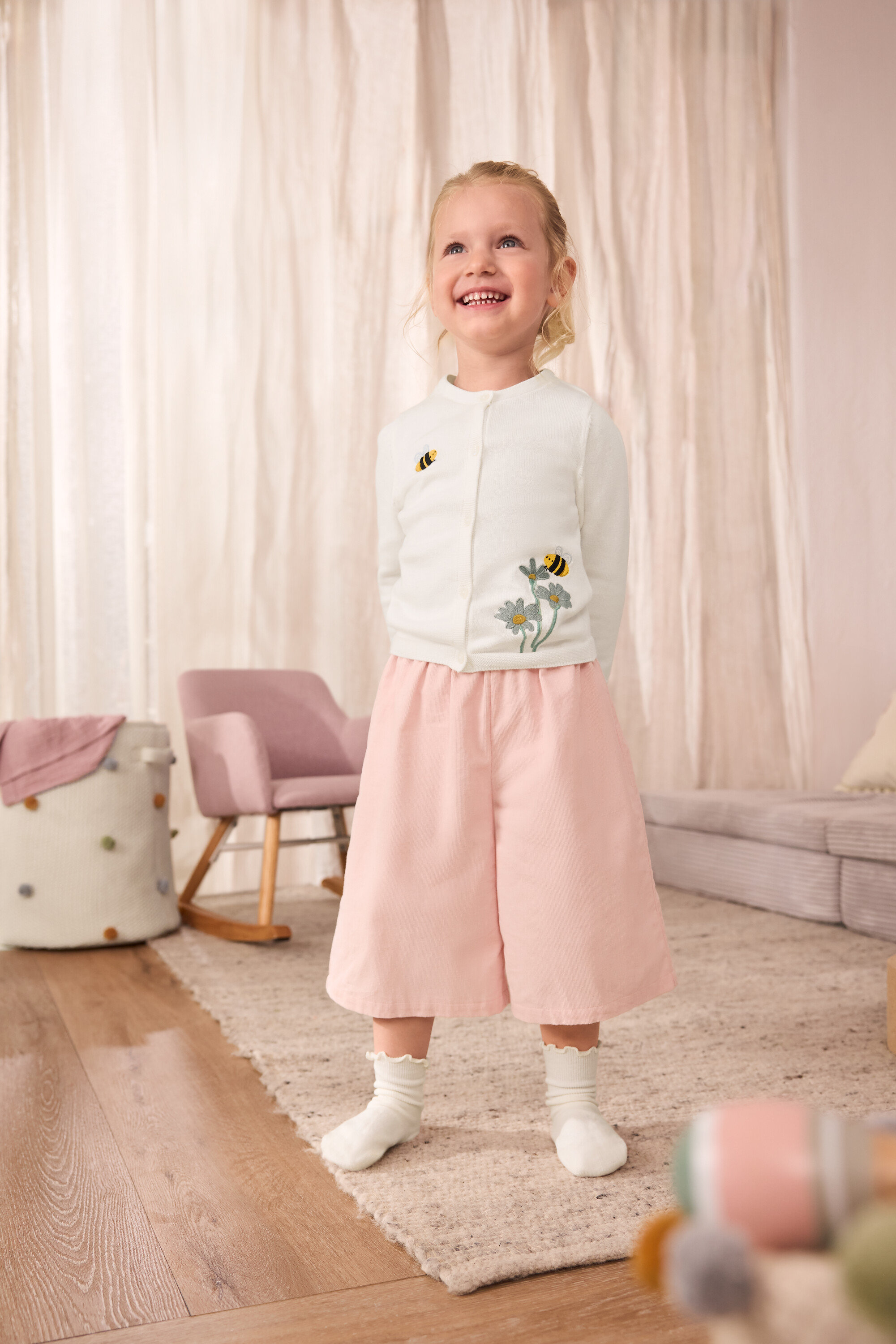 Usmievavé dievča má na sebe detský pletený sveter s vyšitými včielkami, detské menčestrové culottes nohavice a detské ponožky s volánikmi, 5 párov.
