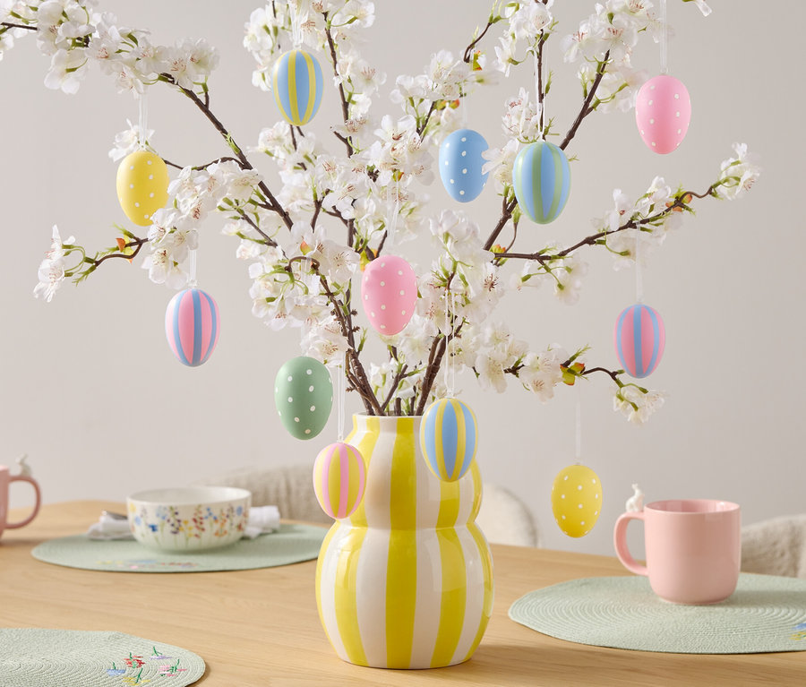 Dekoratívny stôl s veľkonočnými vajíčkami na kvitnúcich konároch v žlto-bielej pruhovanej váze, ružové hrnčeky a miska.