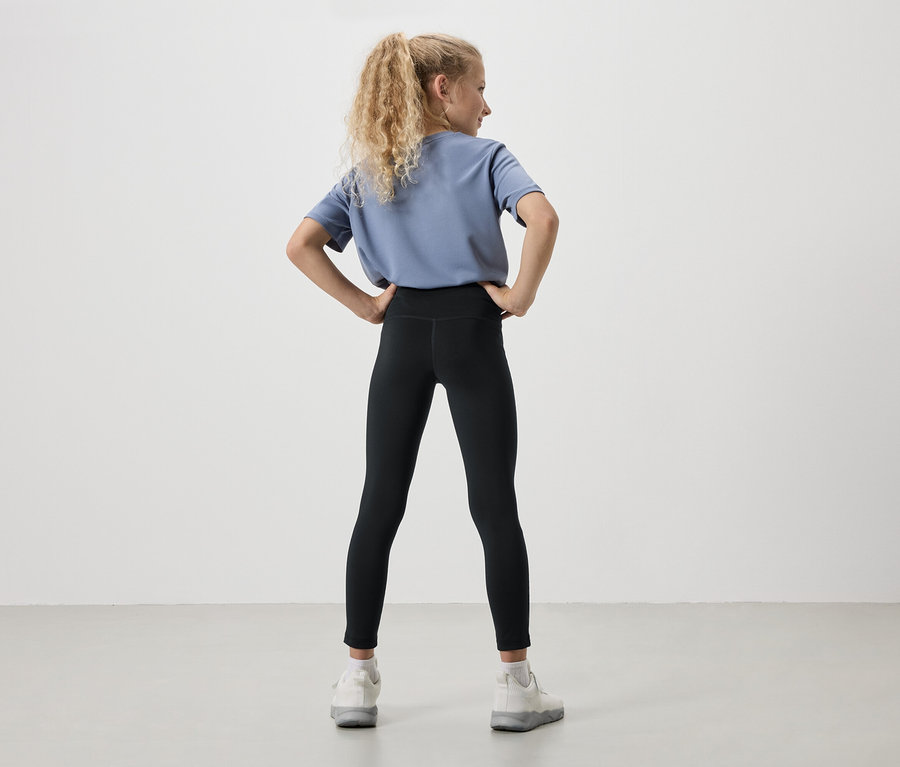 Pohľad zozadu na mladé dievča s blond kučeravými vlasmi, oblečené v modrom tričku, čiernych legínach a bielych teniskách, s rukami vbok.