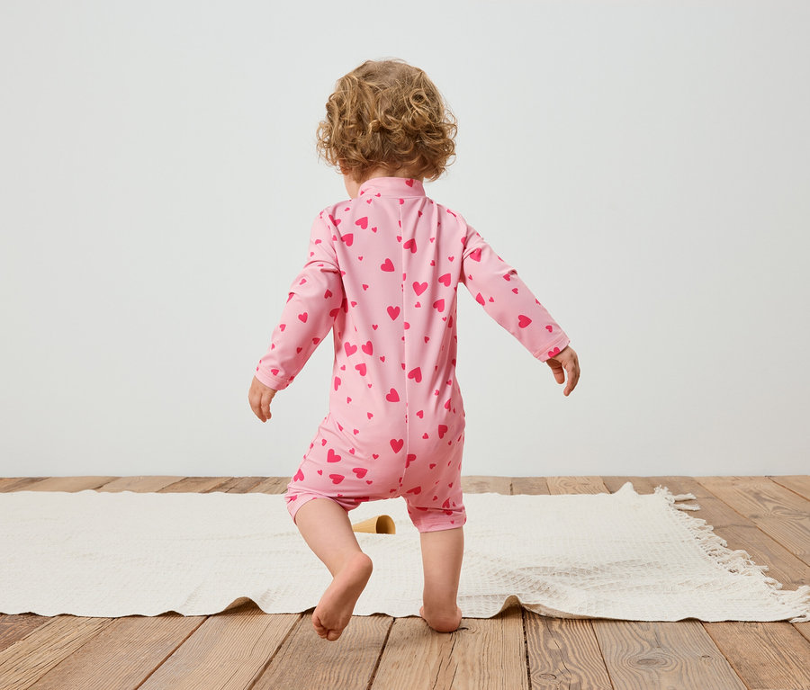 Dieťa zozadu, má na sebe ružový overal so vzorom srdca, stojí na koberci.