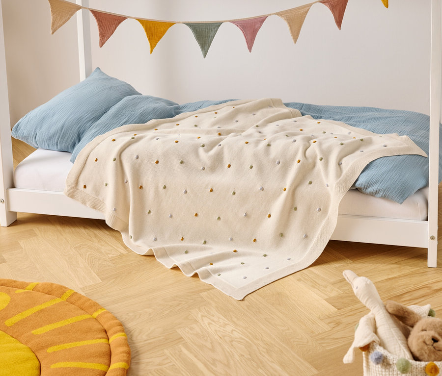 Detská posteľ s modrými vankúšmi a krémovou prikrývkou s farebnými bodkami stojí na drevenej podlahe. Nad posteľou visí farebná vlajková girlanda.