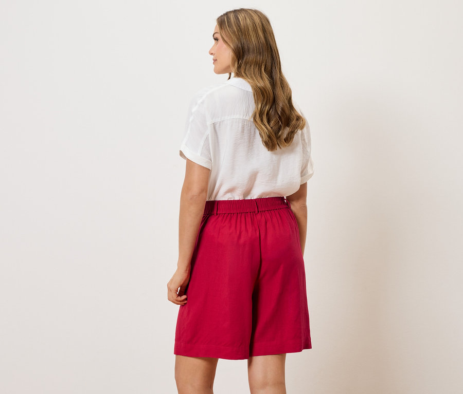 Pohľad zozadu na ženu s vlnitými vlasmi, oblečenú v bielej blúzke a červených šortkách.