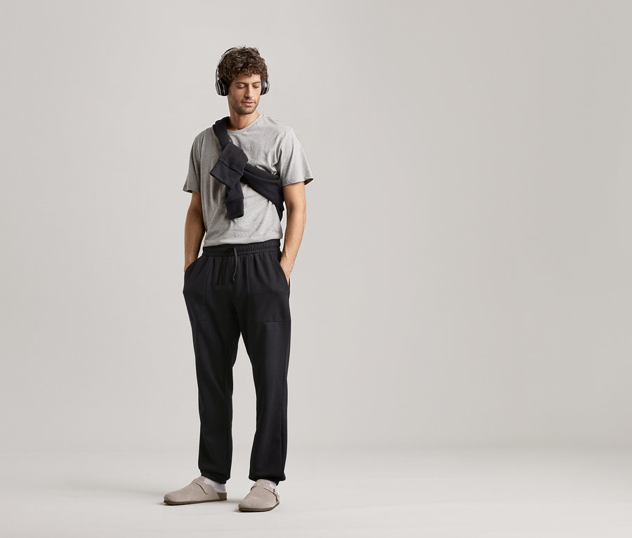 Muž so slúchadlami, sivým tričkom a čiernymi teplákmi na neutrálnom pozadí.