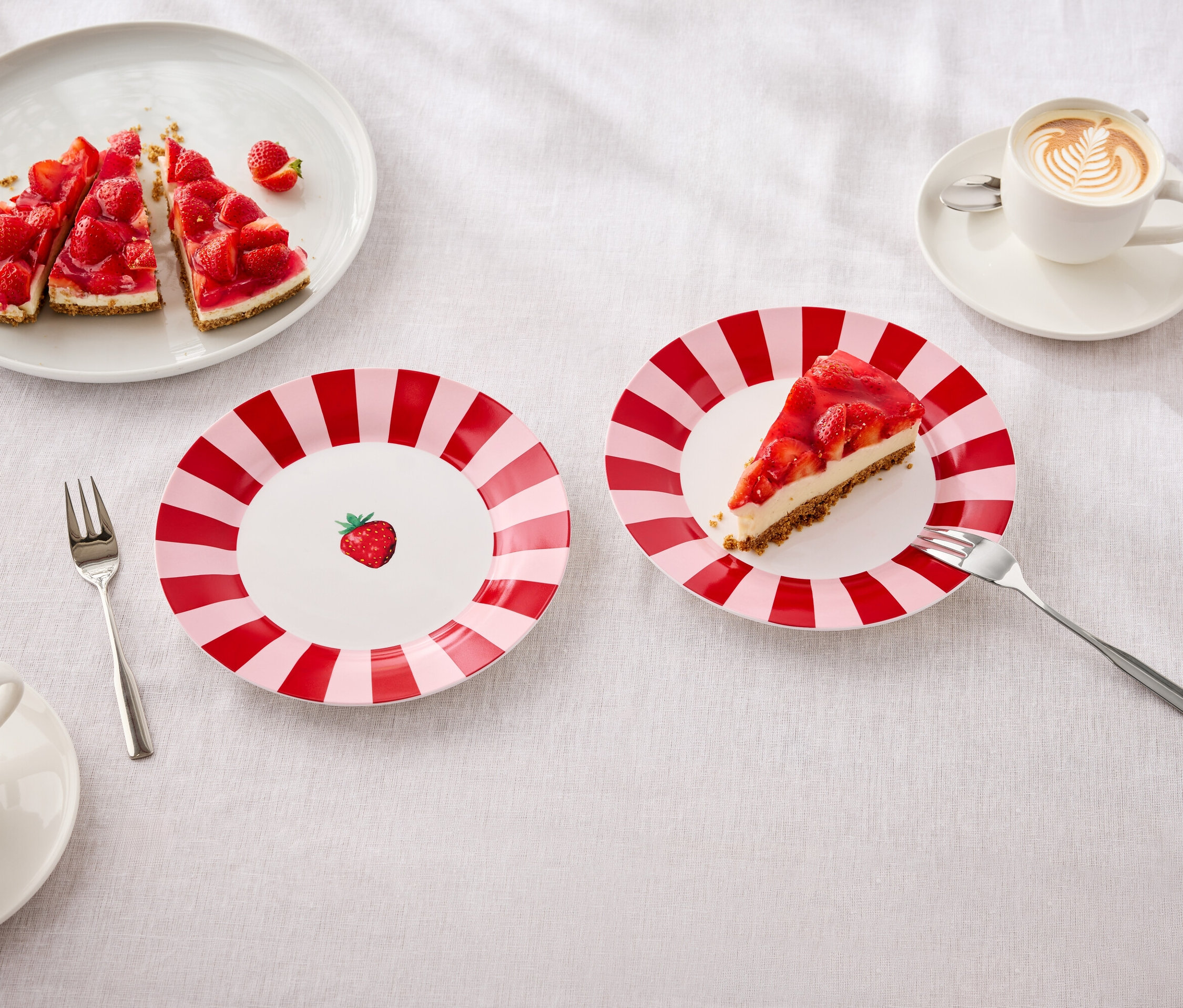 Na stole sú taniere s jahodovým koláčom, vidličky a šálky s latte art.