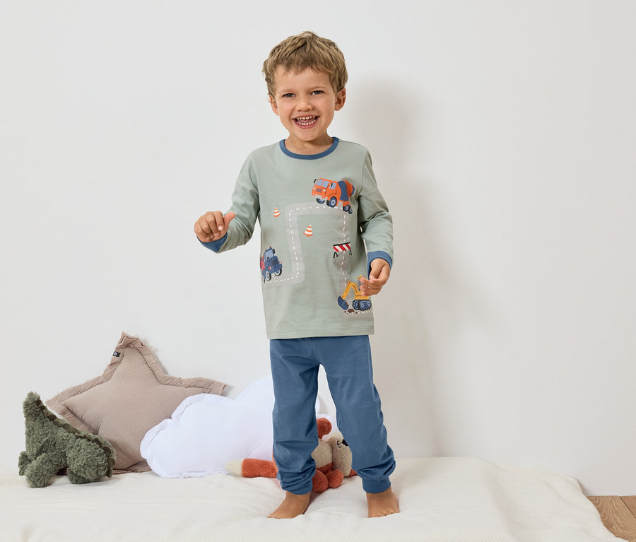 Usmievavý chlapec stojí bosý na bielom povrchu a má na sebe sivé pyžamo s modrými manžetami a motívom bagra a miešačky betónu.