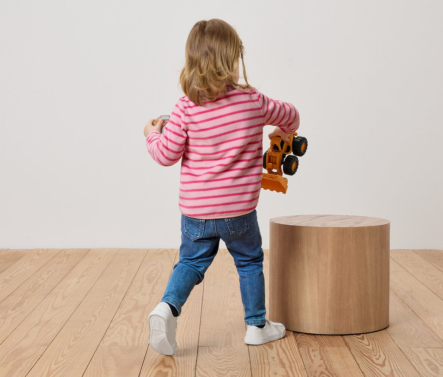 Dieťa s blond vlasmi stojí chrbtom k divákovi a drží v ruke hračku bagra.