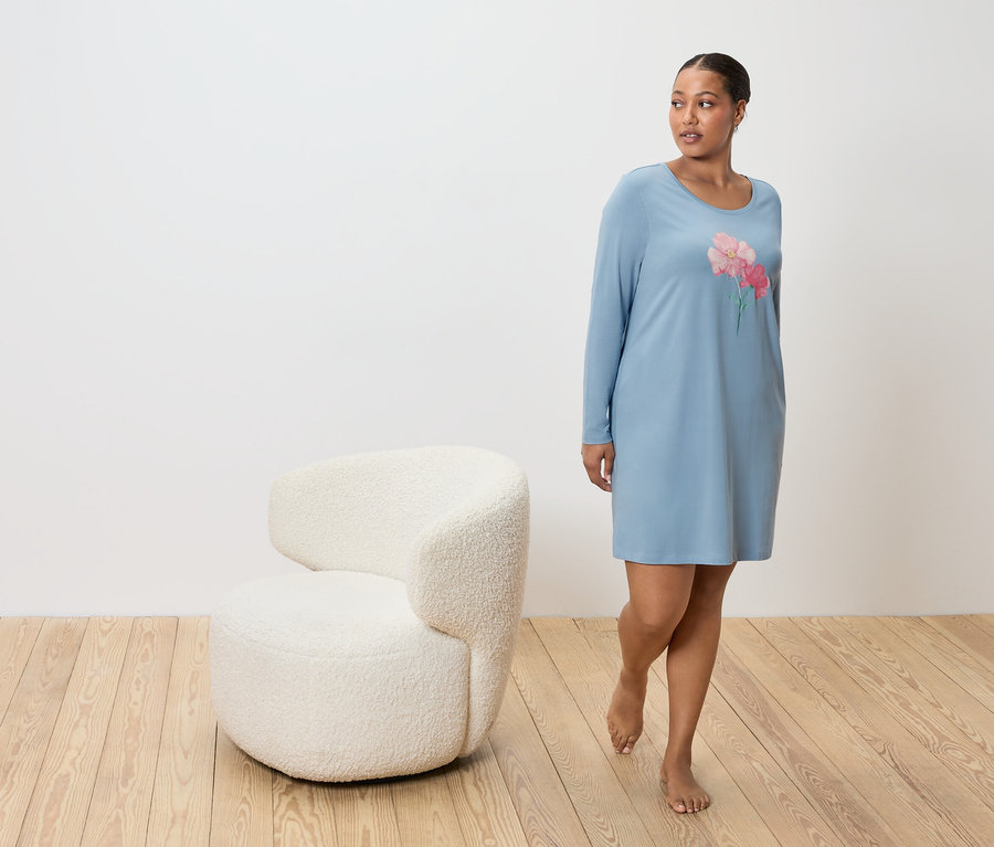 Žena v modrej nočnej košeli s kvetinovým vzorom, vedľa bieleho kresla na drevenej podlahe.