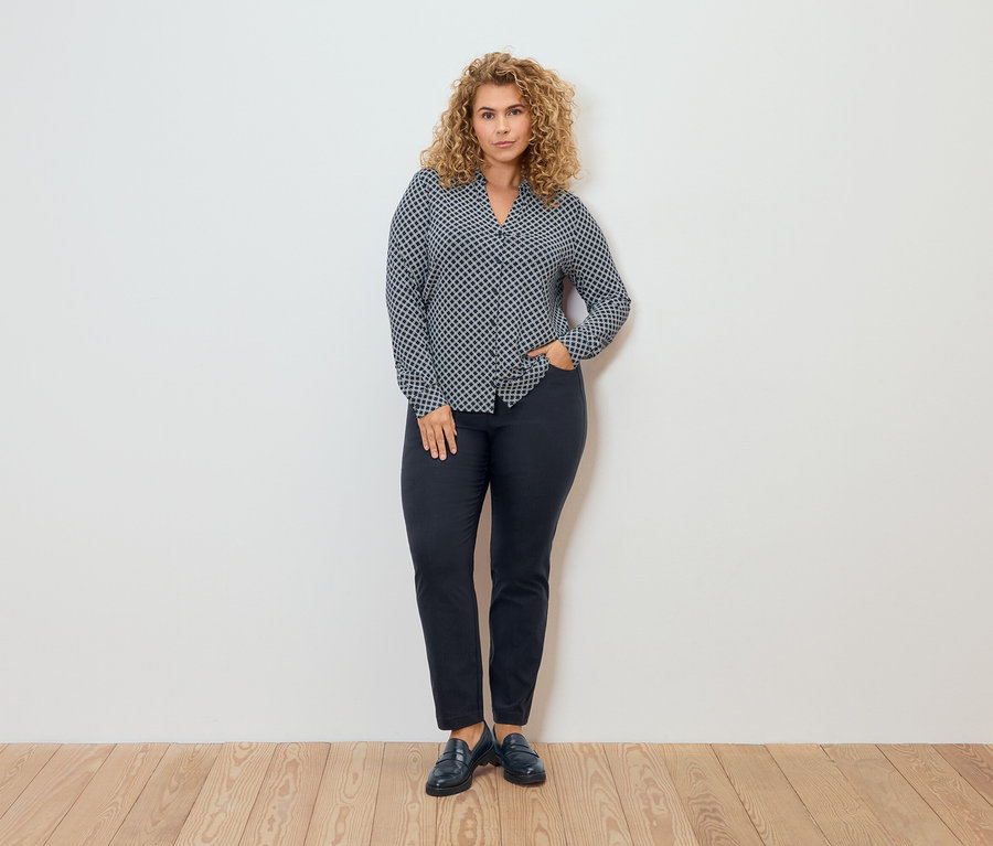 Žena v blúzke so vzorom a nohaviciach z bengalínu pózuje pred stenou.