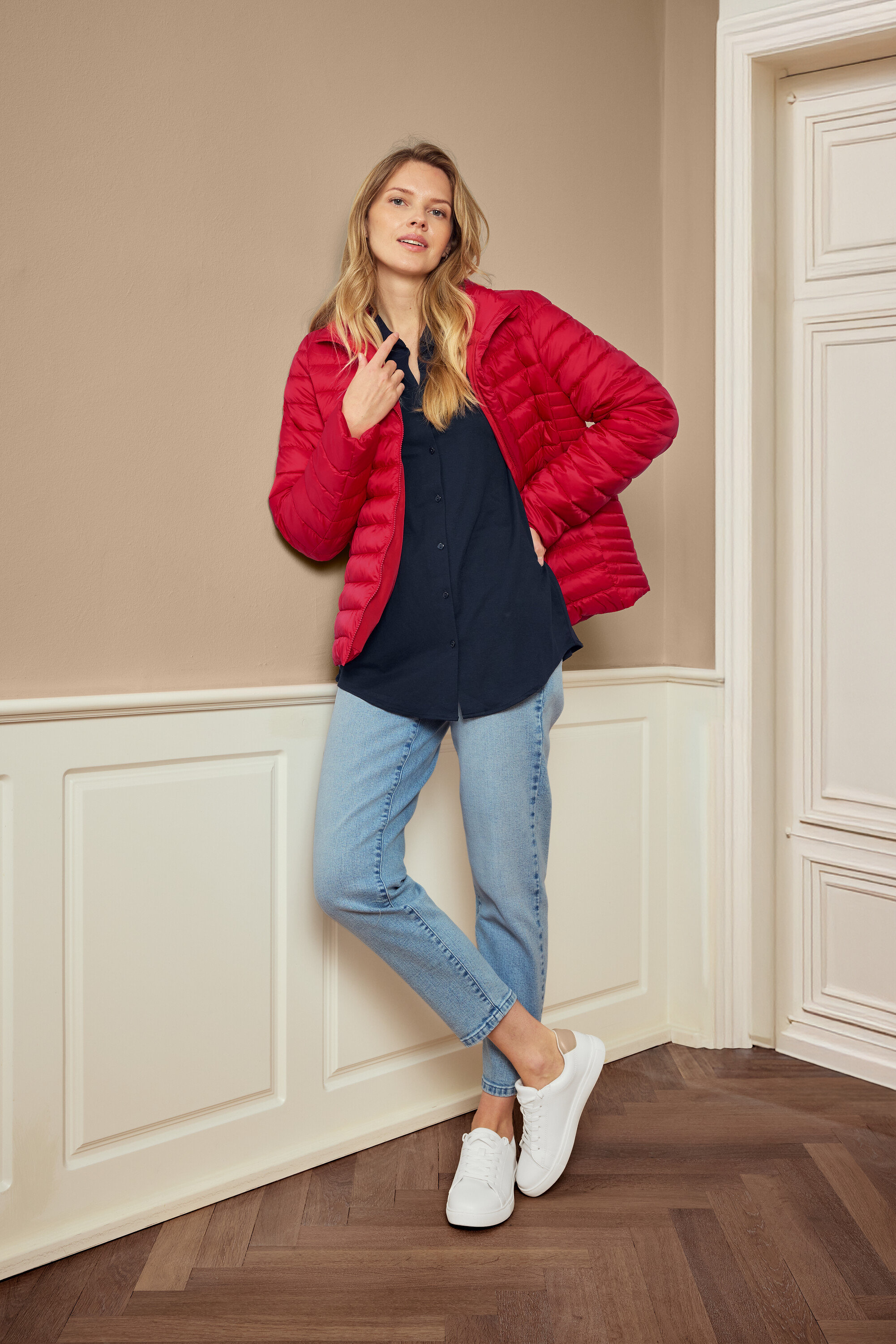 Žena pózuje v červenej bunde, modrej košeli, džínsoch a bielych teniskách, nosí náušnice 925 Silber Creolen Hexagon.