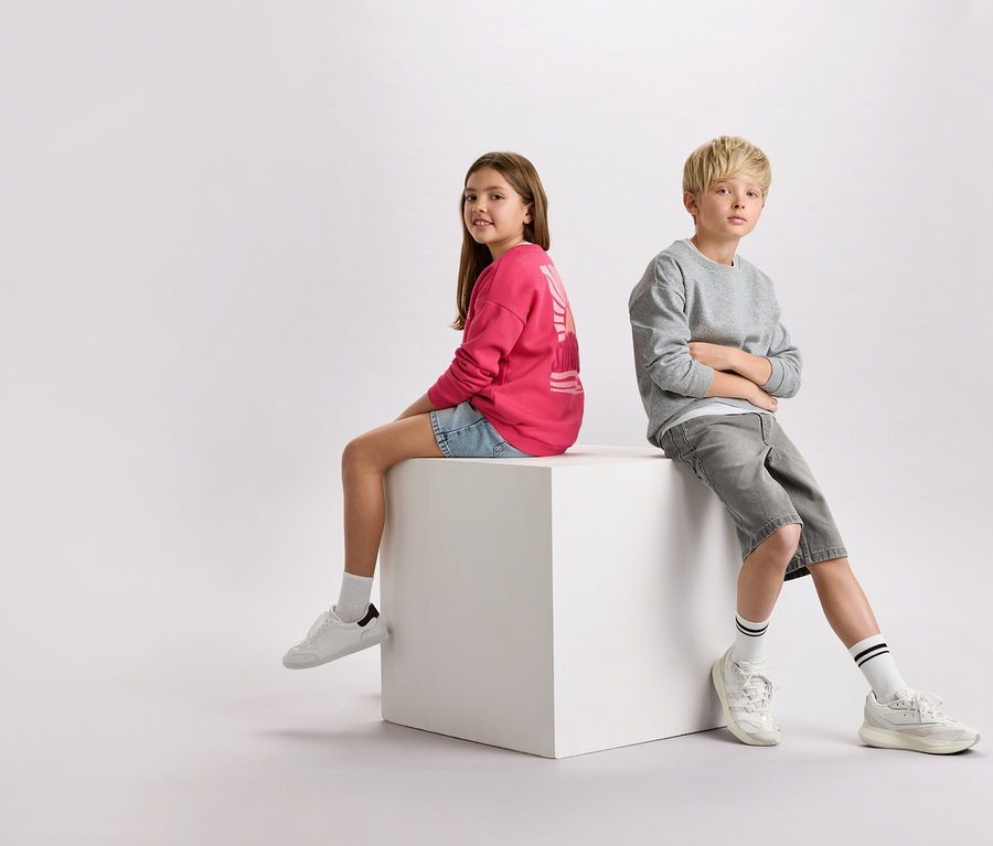 Dievča s hnedými vlasmi v ružovej mikine a džínsových šortkách sedí vedľa chlapca s blond vlasmi v sivej mikine a šortkách na bielej kocke.