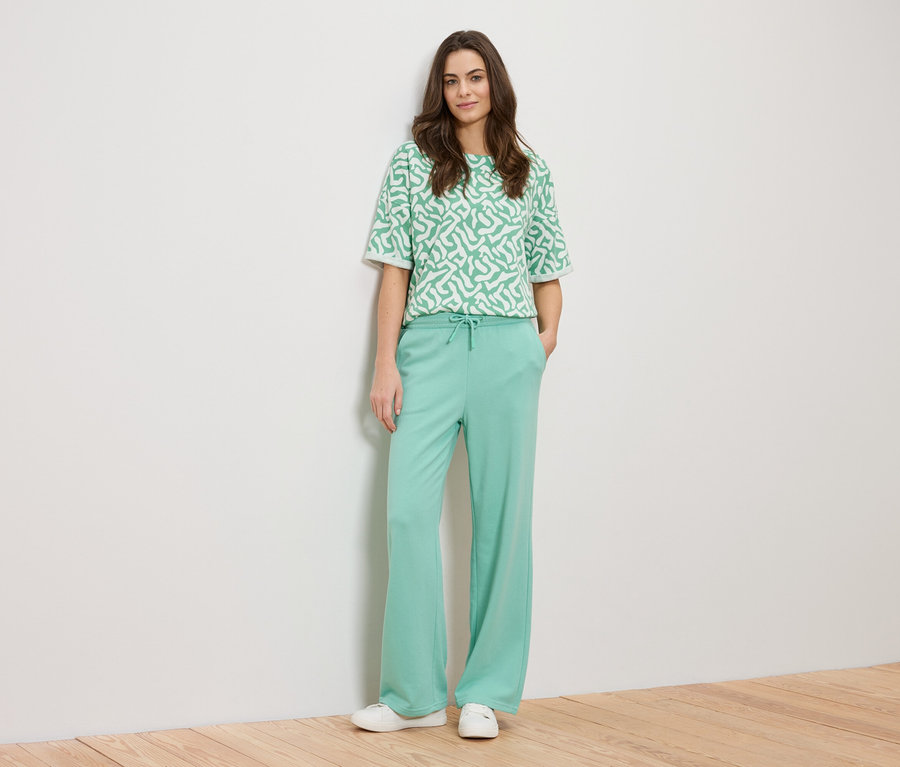 Žena sa opiera o stenu a má na sebe zelený vzorovaný top a zelené nohavice.