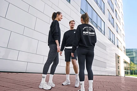 Traja ľudia, dve ženy a jeden muž, stoja usmiati a rozprávajú sa. Všetci majú na sebe čierne mikiny a biele unisex ponožky Coffee Crew. Muž má na sebe čierne športové šortky. Jedna žena má sivo-čierne športové legíny »ActivePower«, druhá čierne športové legíny »ActivePower«.