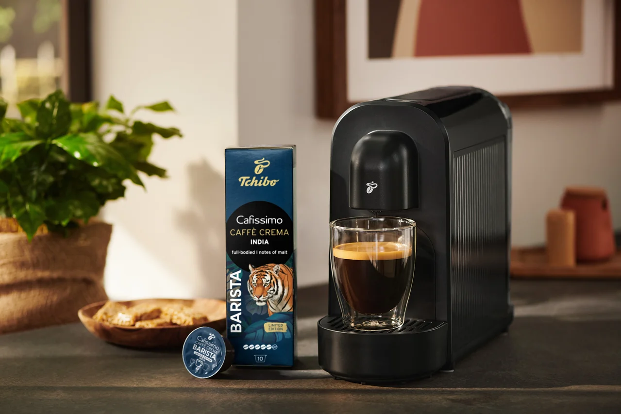 Čierny kávovar Tchibo Cafissimo so šálkou kávy, vedľa balenie kávových kapsúl Tchibo Cafissimo Caffè Crema India a jedna kapsula.