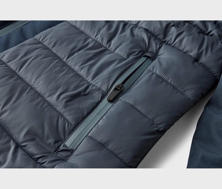 Detail tmavomodrej prešívanej bundy s horizontálnym prešívaním a diagonálnym vreckom na zips. Zips má čierny jazdec a svetlejšiu modrú pásku.