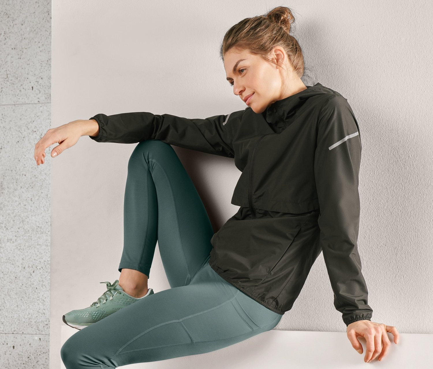 Žena sedí na okraji a má na sebe čiernu vetruvzdornú bundu na behanie, zelené legíny a tenisky.