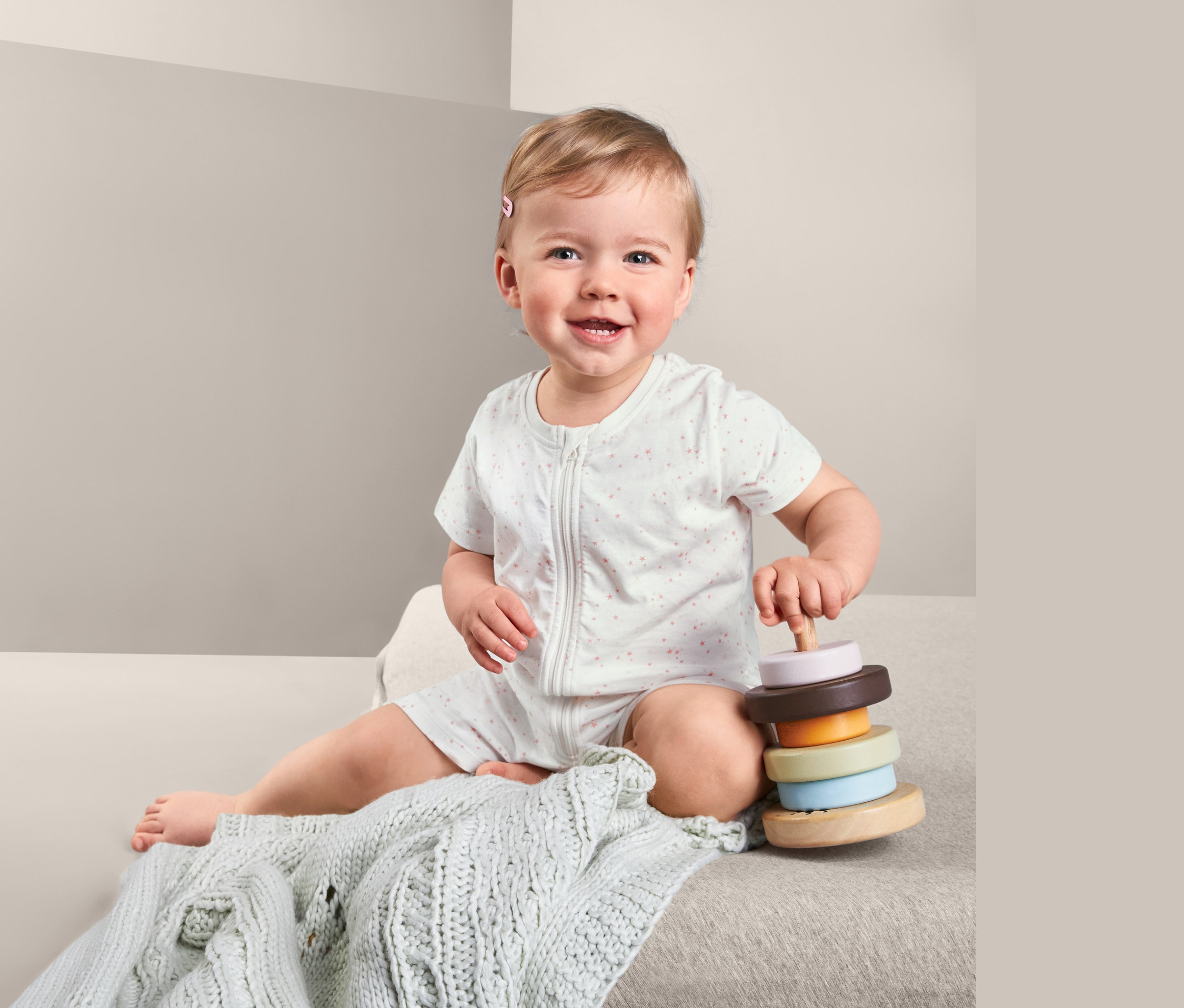 Usmievajúce sa dieťa sedí a hrá sa s hračkou s krúžkami. Má na sebe krátke pyžamá, 2 ks, ružovo-biele.